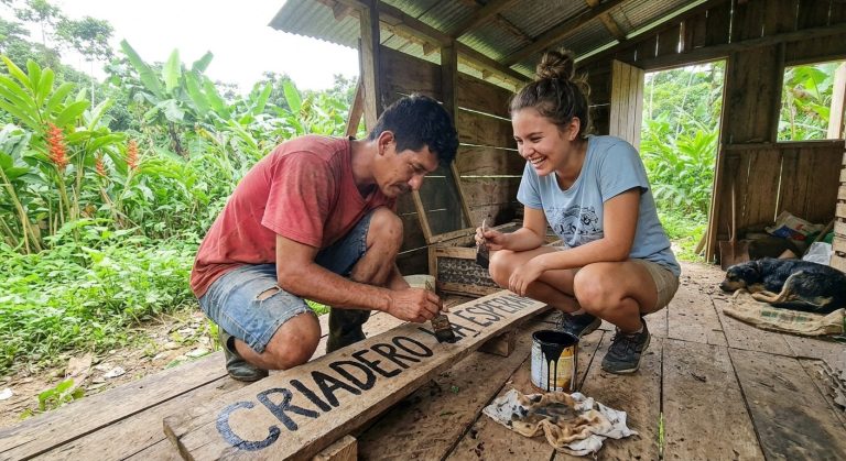 Two people crouched on the farm floor painting the wooden CRIADERO sign